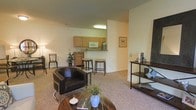 This single-floor residence features an open-concept design that seamlessly integrates the living area and kitchen, creating a spacious and cohesive layout. The living area is anchored by plush carpeting and accommodates a comfortable seating arrangement, including a leather accent chair and a round wooden coffee table, contributing to a relaxed atmosphere. A console table along one wall provides additional display and storage space.

The kitchen is positioned at the rear of the open layout and is outfitted with light wood cabinetry, a white microwave, and standard appliances. A raised breakfast bar with two bar stools serves as both a functional dining surface and a visual divider between the kitchen and living space. Overhead lighting illuminates the kitchen area, ensuring a well-lit workspace.

The neutral color palette throughout — with warm beige walls and complementary carpeting — lends the space a calm, understated aesthetic. Decorative touches such as potted plants and framed wall art add character to the interior. The open flow of the floor plan enhances the sense of space, making the residence feel airy and versatile.