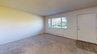 Located at 11020 SE Kent Kangley Rd in Kent, Washington, this one-bedroom, one-bathroom unit offers a practical and efficiently designed single-floor layout. The living room provides a comfortable, open feel with soft carpeted flooring, a textured ceiling, and a wall sconce for ambient lighting. It connects seamlessly to the kitchen, which features white cabinetry, a white range, a refrigerator, and a built-in countertop passthrough — keeping the space feel open and connected. The bedroom is a well-proportioned retreat with plush carpeting, a wide window that brings in natural light and outdoor views of surrounding greenery, and a baseboard heater for year-round comfort. A second carpeted room — visible from the bedroom — leads directly to the bathroom and provides additional flexible space. The bathroom is clean and functional, equipped with a full-size soaking tub and shower combo featuring built-in shelving, chrome fixtures, and a shower curtain rod. Throughout the unit, neutral warm tones, textured ceilings, and carpeted floors contribute to a consistent and understated aesthetic. The unit also includes an electrical panel accessible from within the living space.