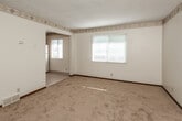 This single-floor residence offers a practical and straightforward layout designed for comfortable everyday living. The living area is generously sized, featuring warm beige carpeting, neutral cream walls, a textured ceiling, and a decorative diamond-patterned border trim. Natural light enters through horizontal-blinded windows, creating a bright and open atmosphere. A floor-plan overview reveals a well-organized open flow between the living space and kitchen area.

The kitchen is equipped with rich, dark wood cabinetry featuring detailed raised-panel doors and ample upper and lower storage. White appliances, including a coil-burner electric range, refrigerator, and double-basin sink with a light-colored countertop, provide full functionality. Light ceramic tile flooring ties the kitchen together cleanly.

The bedrooms are carpeted and feature the same neutral tones and decorative border trim found throughout the unit. At least one bedroom includes a built-in closet with a hanging rod, offering practical storage. Each room benefits from natural light via windows with blinds.

The bathroom is tiled underfoot and includes a vanity with a marble-patterned countertop, a toilet, and a shower-tub combination enclosed by a curtain. Overall, the space presents a clean, functional interior well-suited for residential use.