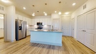 This single-floor residence showcases a thoughtfully designed open-concept layout with consistent, modern finishes throughout. The kitchen serves as the centerpiece of the living area, anchored by a generously sized island with a speckled granite countertop, an undermount sink, and a gold-toned faucet. White shaker-style cabinetry lines the walls, paired with a subtle tile backsplash and matching granite countertops. Stainless steel appliances, including a French door refrigerator with an ice and water dispenser, an electric range, and an over-the-range microwave, complete the functional cooking space. A trio of globe pendant lights with gold accents hangs above the island, adding a decorative touch. Light wood-look flooring flows throughout the main living areas, lending a clean and cohesive aesthetic. The bedrooms are finished with plush neutral carpeting and feature ceiling fans, bright windows with slatted blinds, and views of the surrounding area. Two bathrooms serve the unit: one with a tub-shower combination and a curved rod, and another with a single vanity topped with granite, white cabinetry, gold hardware, and a large mirror. The overall ambiance is bright, clean, and move-in ready.