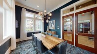 This single-floor space makes a strong design statement through its carefully curated aesthetic and functional layout. The focal point of the room is a substantial live-edge natural wood dining or conference table, surrounded by tufted charcoal leather chairs that balance sophistication with comfort. Overhead, a brass sputnik-style chandelier with globe bulbs serves as a dramatic centerpiece, casting warm light throughout the space.

Deep navy walls provide a bold, refined backdrop, contrasted by crisp white crown molding and window trim that frame an array of large windows on two walls. These generously sized windows flood the room with natural light while offering views of the surrounding neighborhood, enhancing the sense of openness. Sheer white drapery softens the light without obstructing the outlook.

Two wall-mounted flat-screen televisions are positioned on opposing navy walls, making the space well-suited for presentations or group viewings. A patterned area rug in dark, moody tones anchors the seating area and adds textural depth underfoot. A wooden sideboard with decorative objects sits against the accent wall alongside curated artwork.

Entry is through elegant glass-paneled wood doors that connect this room to a broader hardwood-floored common area, reinforcing its character as a communal gathering or private meeting space.