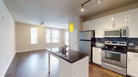 This single-floor residence presents a thoughtfully designed open-concept layout that seamlessly integrates the kitchen, dining, and living areas. The kitchen is a standout feature, showcasing white shaker-style cabinetry extending to the ceiling, dark granite countertops, and a striking mosaic tile backsplash in blue and grey tones. A central island with a dark countertop serves as both a workspace and casual dining area, accented by a vibrant yellow pendant light. Stainless steel appliances, including a refrigerator, range, microwave, and dishwasher, complete the functional kitchen setup. Rich, dark hardwood-style flooring runs throughout the main living areas, adding warmth and contrast to the neutral grey walls. The living area benefits from multiple large windows fitted with blinds, allowing generous natural light to fill the space. Track lighting installed on the ceiling provides additional illumination. The bedroom is generously sized, featuring plush carpet flooring, a ceiling fan, and corner windows that bring in abundant natural light and outdoor views. The overall aesthetic is clean, modern, and cohesive, with a neutral palette throughout.