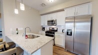 This thoughtfully designed one-bedroom, one-bathroom residence with a den is located in Livonia, MI, offering a comfortable and modern single-floor layout. The kitchen is a standout feature, showcasing crisp white shaker cabinetry, light granite countertops with subtle speckled detailing, a double-basin stainless steel sink, and a full suite of stainless steel appliances including a gas range, over-the-range microwave, and a French door refrigerator with an in-door ice and water dispenser. Wood-look flooring runs throughout the kitchen and main living areas, lending warmth and visual continuity to the space. The open layout connects the kitchen seamlessly to the living and dining areas, creating an inviting flow ideal for everyday living and entertaining. The bedroom is generously proportioned, featuring plush carpeting, a tray ceiling detail, and a bay-style window arrangement that fills the room with natural light and offers pleasant outdoor views. The full bathroom is finished in clean, neutral tones with a soaking tub and tiled surround with built-in corner shelving. The additional den provides flexible space that can serve as a home office, reading nook, or guest area. Neutral wall tones throughout create a calm, cohesive ambiance.