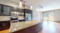 This single-floor residence offers a thoughtfully designed layout that balances functional living spaces with contemporary finishes throughout. The kitchen serves as a central highlight, showcasing rich dark cabinetry with sleek hardware, granite countertops with warm gold and beige tones, and a full suite of stainless steel appliances including a range, built-in microwave, refrigerator with ice dispenser, and dishwasher. A generously sized granite-topped island provides additional prep space and casual seating. Pendant lighting and rail-mounted track lights illuminate the kitchen and flow into the adjoining living area, which features warm hardwood flooring and a sliding glass door that invites natural light and outdoor views. A dedicated in-unit laundry closet with a stacked washer and dryer adds everyday convenience. The bedroom offers plush carpeting, elevated ceilings, and a bright window with views of the surrounding neighborhood. The full bathroom is well-equipped with dark cabinetry, granite countertops, a large vanity mirror, tile flooring, and a separate water closet, providing a clean and functional layout throughout.