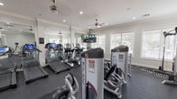 This single-story fitness center in Humble, TX is thoughtfully designed to support a full range of workout routines. The space features a clean, modern aesthetic with white walls, crown molding, and recessed lighting complemented by multiple ceiling fans that maintain air circulation throughout. Rubber flooring covers the entire workout area, providing both safety and durability underfoot.

The cardio zone is well-appointed with a row of treadmills, elliptical trainers, and cycling bikes, all from recognized equipment brands. Large wall mirrors run along one side of the room, visually expanding the space and allowing users to monitor their form. Wall-mounted flat-screen televisions are positioned for easy viewing from the cardio equipment.

The strength training area includes Body-Solid selectorized machines targeting various muscle groups, adjustable weight benches, a cable/pulley multi-station unit, and a dumbbell rack. A dedicated stretching and free-weight zone offers open floor space for functional training.

Natural light filters in through multiple windows fitted with horizontal blinds, offering views of the surrounding outdoor greenery. A glass partition and interior windows connect the fitness center to an adjacent room, adding openness to the overall layout. The space is also equipped with security cameras and clearly marked emergency exits.