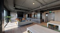 This single-floor space showcases a sophisticated industrial-modern aesthetic throughout. Exposed ductwork and services run across dark ceilings, complemented by pendant and track lighting that creates a warm, layered ambiance. Light hardwood flooring flows consistently through all areas, unifying the open layout.

The lounge area serves as a central gathering point, furnished with a neutral sofa, tufted navy ottoman, and cognac leather armchairs arranged around an artistic chalkboard feature wall with botanical illustrations. A wall-mounted television anchors this zone. Floor-to-ceiling sliding glass doors open directly onto an outdoor terrace, flooding the space with natural light and offering sweeping city views.

The kitchen features dark countertops, warm wood cabinetry, dual sinks, and pendant lighting, with a striking black-and-gold marble-patterned partition wall creating visual separation from adjacent areas. Nearby, multiple dining tables with green upholstered chairs accommodate both casual and formal gatherings, also benefiting from direct terrace access and abundant natural light.

A more enclosed meeting or dining area provides a quieter setting, with light wood paneling, frosted glass entry doors, and a clean, understated finish. Throughout the space, the interplay of dark architectural elements, warm wood tones, curated furnishings, and outdoor connectivity creates a refined yet functional communal environment.