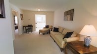 This single-floor space offers a welcoming and functional layout designed for comfortable everyday living. The living area serves as the heart of the home, featuring plush carpeted flooring and neutral-toned walls that create a warm, inviting atmosphere. The space is furnished with a generous sofa and matching armchair, both in a soft cream upholstery, accompanied by an upholstered ottoman that adds versatility to the seating arrangement. Decorative artwork adorns the walls, adding subtle character to the room. A compact dining area is positioned to one side, complete with a dark-finished table and chairs, making efficient use of the available space. Natural light filters in from the adjacent kitchen, which is visible through an open doorway and features white cabinetry, light countertops, and a window that allows additional brightness into the area. The open sightline between the living area, dining space, and kitchen enhances the sense of flow and connectivity throughout the home. Overall, the space presents a clean, understated aesthetic with a practical and comfortable design well-suited for day-to-day living.