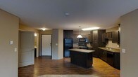 This single-floor residence offers a well-designed open-concept layout that seamlessly connects the kitchen and living areas. The kitchen is a standout feature, showcasing rich, dark wood cabinetry that extends to the upper walls, providing ample storage space. A central island anchors the kitchen, offering additional counter space and serving as a natural gathering point. Black appliances, including a refrigerator, microwave, and range, complement the dark cabinetry for a cohesive aesthetic. Warm pendant lighting hangs above the island, adding both function and style to the space. The kitchen transitions effortlessly into the open living area, all unified by warm hardwood-style flooring that runs throughout. Neutral-toned walls provide a clean backdrop that balances the darker furnishings and cabinetry. A hallway off the main living area leads to additional rooms, including what appears to be a bathroom with its own lighting. The front entry features a solid white door, offering a bright contrast to the darker interior palette. Overall, the space presents a warm, inviting ambiance with a practical and stylish design suited for everyday living.