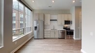 This single-floor unit showcases a thoughtfully designed open-concept layout with a contemporary aesthetic. The kitchen is a standout feature, equipped with stainless steel appliances including a refrigerator with ice maker, electric range, built-in microwave, and dishwasher. Light wood-tone cabinetry with brushed metal hardware pairs with crisp white quartz countertops and a subway tile backsplash in a vertical stacked pattern, creating a clean, modern look. A stainless steel undermount sink and recessed lighting complete the space.

The living area flows naturally from the kitchen and is bathed in natural light from large floor-to-ceiling windows, offering views of the surrounding building complex and community amenities. Light wood-look flooring runs throughout the main living spaces, adding warmth and continuity to the design.

The bathroom maintains the same neutral, modern palette with warm beige walls, wood-look flooring, a white vanity with an undermount sink, and a framed mirror. A glass-enclosed shower with tiled walls and built-in shelving provides functional storage. The overall ambiance of the unit is bright, clean, and contemporary, ideal for everyday living.