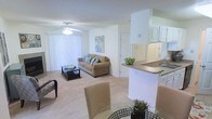 This single-story residence showcases an open-concept floor plan that seamlessly integrates the living, dining, and kitchen areas into one cohesive space. The living room is finished with wall-to-wall carpeting and features a ceiling fan with a light fixture, providing both comfort and airflow. Large vertical blinds covering a sliding glass door allow natural light to filter into the room while offering privacy control. A neutral color palette of whites and soft beiges carries throughout, creating a bright and airy ambiance.

The kitchen is positioned adjacent to the living area, separated by a peninsula counter with a dark laminate surface that doubles as a breakfast bar. White cabinetry lines the kitchen walls, complemented by a stainless steel sink. The pass-through design of the kitchen enhances the sense of openness and maintains visual connectivity with the rest of the living space.

The dining area, situated in the foreground, accommodates a round table with seating, completing the functional layout. A textured popcorn ceiling is consistent throughout, and interior doors provide access to additional rooms beyond the main living area.