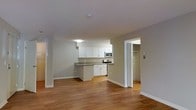 This single-floor space offers a thoughtfully designed open-concept layout that seamlessly connects the living area and kitchen, creating a sense of spaciousness and easy flow throughout. The kitchen is well-appointed with crisp white cabinetry, a neutral countertop, and stainless steel appliances including a built-in microwave and range, providing both style and functionality. The open layout allows the kitchen to serve as a natural focal point while remaining connected to the broader living space.

Warm hardwood-style flooring runs throughout the main area, adding a welcoming tone that complements the neutral gray and white color palette of the walls and trim. Large white baseboards frame the space cleanly, contributing to a polished and well-maintained appearance. The ceiling is smooth and bright, keeping the interior feeling open and airy.

A doorway visible from the main living area leads to what appears to be a generously sized walk-in closet, complete with shelving and hanging rods, offering practical storage solutions. The overall ambiance of the space is clean, modern, and move-in ready, making it a versatile and comfortable environment suited to a variety of lifestyles.