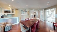 This thoughtfully designed single-floor space combines modern sophistication with warm, rustic character. The main gathering area is anchored by rich, deep-toned hardwood floors that flow throughout, creating a cohesive and inviting atmosphere. A standout stone-surround fireplace serves as a focal point, complemented by recessed lighting that adds warmth and dimension to the room. A sturdy wood dining table surrounded by bold red leather and patterned chairs adds a vibrant pop of color, while a large multicolored reclaimed wood art piece adorns the wall, lending an artistic, eclectic touch.

Distinctive frosted glass panel doors with geometric cutouts provide a stylish division between spaces while allowing light to filter through, maintaining an open and airy feel. The wet bar area is well-appointed with granite countertops, a stainless steel sink, white shaker-style cabinetry, open shelving, and a built-in beverage refrigerator — ideal for entertaining. Tile flooring in a herringbone pattern transitions seamlessly into this functional zone, adding texture and visual interest.

Large windows and glass doors invite natural light and offer views of the surrounding greenery. The overall ambiance is polished yet approachable, blending contemporary finishes with warm accents for a space that is both functional and visually compelling.