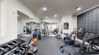This fitness center at 11925 Jones Rd in Houston, Texas is a thoughtfully designed, single-floor facility that balances functionality with a polished aesthetic. The space features a neutral white color palette accented by a distinctive dark plaid feature wall, adding a stylish, boutique-gym feel to the environment. Recessed lighting throughout ensures the space is well-lit, while crown molding along the ceiling lends a refined architectural touch. Wall-mounted industrial fans provide additional airflow across the workout floor.

The cardio section is well-appointed with multiple Echelon treadmills and stationary bikes, each equipped with integrated digital screens for interactive workout experiences. A rowing machine rounds out the cardio offerings. The strength training area features a multi-station cable machine, a flat bench, a rack of hex dumbbells spanning a wide weight range, colorful medicine balls, and a dedicated medicine ball storage rack. Full-length mirrors along one wall amplify the sense of space and allow users to monitor their form during exercise.

Natural light enters through windows facing the exterior, offering views of the surrounding property. A glass-paneled door provides direct outdoor access. A posted Fitness Center Rules sign reflects attentive facility management. The carpet flooring throughout is practical and durable, suited for a high-use fitness environment.