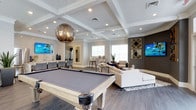 This sophisticated single-floor amenity space blends refined design with leisure-focused functionality. At its heart sits a full-size billiards table with a light wood frame, anchored beneath a striking smoked-glass globe pendant light that serves as a dramatic focal point. The coffered ceiling with recessed lighting adds architectural elegance and a sense of volume to the open layout.

The space flows seamlessly into a comfortable lounge area furnished with plush cream-toned sectional seating, accented by colorful tropical artwork and bold geometric wall décor. Natural light pours in through large windows, brightening the neutral color palette of soft grays and whites.

Along one wall, a wet bar with counter seating and a mosaic tile backsplash provides a stylish entertaining station, complete with bar-height chairs and decorative bottles. Dark hardwood-style flooring runs throughout, grounding the space with warmth and contrast against the lighter furnishings.

Thoughtfully curated decorative touches — including tropical-themed canvas prints and metallic accent pieces — give the space a cohesive, resort-inspired aesthetic. Whether used for social gatherings or casual relaxation, this well-appointed space offers an inviting and upscale atmosphere designed for both entertainment and comfort.