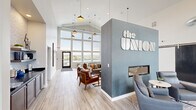 The Union Clubhouse, located on Berky Drive in Box Elder, South Dakota, is a thoughtfully designed community gathering space that blends modern industrial aesthetics with warm, inviting interiors. The centerpiece of the space is a dramatic double-height wall of floor-to-ceiling windows that floods the interior with natural light and offers sweeping outdoor views, including a glimpse of the community pool area beyond.

A signature textured blue-grey accent wall bearing "the Union" logo in bold three-dimensional lettering anchors the entrance, which also features a sleek built-in fireplace and a cozy seating nook with contemporary lounge chairs. Exposed ductwork and pendant lighting suspended from the soaring ceilings contribute an industrial-chic character throughout.

The main lounge area is generously appointed with a large sectional sofa, tufted cushions, and mid-century-inspired leather accent chairs arranged around a substantial wooden coffee table. A bar-height counter with seating provides a casual workspace or social perch. Toward the rear, a beverage station and shelving offer convenient amenities for residents. Light wood-look flooring runs throughout, tying together the crisp white walls and varied seating zones into a cohesive, welcoming environment ideal for socializing or relaxing.
