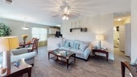 Located at 800 Churchill Downs Dr in Aberdeen, NC, this single-floor residence offers a thoughtfully designed layout that balances comfort and functionality. The open-concept living and dining area is anchored by wood-style flooring, a ceiling fan with light fixture, and windows dressed with layered curtains and blinds that allow ample natural light to fill the space. A peninsula counter connects the living area to the kitchen, which features white cabinetry, light countertops, and black appliances including an electric range and dishwasher. A dedicated dining space accommodates a round table beneath a pendant light fixture. The bedroom includes plush carpeting and neutral wall tones, offering a calm and restful atmosphere. A second bedroom is also present, providing additional flexibility. The primary bathroom is equipped with a wide vanity, a speckled countertop, large mirror, and a tub-shower combination. A half bath includes wood-style flooring and built-in shelving for added storage. Throughout, the interior maintains a cohesive neutral color palette with warm beige and gray tones, contributing to a clean and inviting overall ambiance.