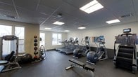 This fitness facility is designed to support a comprehensive workout experience within a single, open floor layout. The space is outfitted with a row of treadmills and elliptical cross-trainers, each equipped with individual digital display screens for performance tracking. A wall-mounted mirror along one side of the room enhances the sense of openness and allows users to monitor their form during exercise. Rubber flooring covers the entire area, providing durability, impact absorption, and safety underfoot. In one corner, a vertical medicine ball rack holds an assortment of weighted balls in varying sizes, accompanied by kettlebells and additional functional training accessories. A weight bench with a barbell is positioned centrally on the floor, offering a dedicated space for strength training. The room benefits from abundant overhead lighting via recessed panel lights set into a drop-tile ceiling, creating a bright and energizing atmosphere. A window near the entrance allows natural light to filter in, adding to the welcoming environment. The clean white walls and efficient use of space give the facility a professional, well-maintained appearance suitable for a wide range of fitness routines.