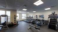 This fitness facility is designed to support a comprehensive workout experience within a single, open floor layout. The space is outfitted with a row of treadmills and elliptical cross-trainers, each equipped with individual digital display screens for performance tracking. A wall-mounted mirror along one side of the room enhances the sense of openness and allows users to monitor their form during exercise. Rubber flooring covers the entire area, providing durability, impact absorption, and safety underfoot. In one corner, a vertical medicine ball rack holds an assortment of weighted balls in varying sizes, accompanied by kettlebells and additional functional training accessories. A weight bench with a barbell is positioned centrally on the floor, offering a dedicated space for strength training. The room benefits from abundant overhead lighting via recessed panel lights set into a drop-tile ceiling, creating a bright and energizing atmosphere. A window near the entrance allows natural light to filter in, adding to the welcoming environment. The clean white walls and efficient use of space give the facility a professional, well-maintained appearance suitable for a wide range of fitness routines.