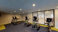 This single-story facility combines sophisticated design with practical amenities across several distinct areas. The main lounge serves as the centerpiece, boasting soaring ceilings that amplify the sense of openness, bathed in natural light that streams through large, dark-framed French doors leading to an outdoor area. Elegant multi-bulb chandeliers illuminate the space, which is furnished with cream-toned tufted sofas and accent chairs in soft neutral tones, creating a refined yet welcoming atmosphere.

A standout feature of the lounge is a floor-to-ceiling stacked stone fireplace, complete with a rustic wooden mantel displaying decorative accents and framed landscape artwork. French doors flanking the fireplace invite natural light and outdoor access, enhancing the airy ambiance. Seating clusters throughout accommodate both intimate conversation and larger gatherings, with plush wingback chairs adding a classic touch.

Adjacent areas include a dining space with a wooden table and upholstered chairs, seamlessly integrated into the lounge environment. The fitness center is outfitted with multiple True-brand treadmills, ellipticals, and stair climbers arranged on patterned carpet, with recessed lighting and a large viewing window providing transparency into the broader facility.