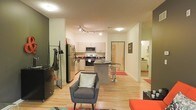 This single-floor unit features an open-concept layout that seamlessly connects the living area to the kitchen, creating a cohesive and efficient use of space. Light hardwood flooring runs throughout, lending warmth and continuity across the entire floor plan.

The kitchen is equipped with stainless steel appliances, including a refrigerator, range, and microwave, complemented by white cabinetry and a dark-topped island with bar-height seating — ideal for casual dining or additional workspace. Track lighting above the kitchen provides focused illumination over the cooking area.

The living space sits directly adjacent, furnished with a contemporary grey armchair and accented by a colorful area rug, adding visual interest to the neutral palette. The walls are painted in soft, neutral tones, contributing to a clean and open ambiance.

Toward the back of the unit, a doorway leads to what appears to be a bathroom, visible with tiled flooring and additional lighting. A clothing rack near the entrance area provides practical storage. Overall, the space offers a modern, functional design with a straightforward, well-connected floor plan.