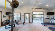 Located at River Ridge in Spokane, WA, this dedicated fitness center offers a thoughtfully designed workout environment spread across a single open floor. The space features striking vaulted ceilings that lend an open, airy atmosphere, complemented by recessed lighting and multiple windows trimmed in rich wood that allow ample natural light to filter throughout. A standout architectural element is the full-width glass-panel garage door, which opens to an adjacent outdoor patio area, seamlessly blending indoor and outdoor space while providing views of the surrounding landscape. The interior is finished with neutral-toned carpet flooring and clean white walls accented by warm wood baseboards and door trim. The fitness area is comprehensively equipped with cardio machines including treadmills and an elliptical, as well as strength training equipment such as a multi-function power rack with resistance bands and free weights, a rowing machine, and adjustable benches. A large wall-mounted mirror enhances the sense of space, while a wall-mounted television provides entertainment during workouts. A secondary glass-pane entry door offers additional access and light. The overall ambiance is clean, functional, and well-lit, making it a versatile and inviting space for a full range of fitness activities.