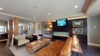 This single-floor amenity space offers a thoughtfully designed environment that blends modern style with practical functionality. The centerpiece is an expansive lounge area featuring rich, dark hardwood flooring, a bold accent wall with a wall-mounted flat-screen TV, a sleek linear gas fireplace, and floating wood shelves with ambient pendant lighting. A generously sized tufted leather sofa anchors the space, complemented by accent chairs and a reclaimed wood coffee table atop a striped area rug. Large windows along one wall flood the lounge with natural light and provide views of lush greenery outdoors, enhancing the open and airy atmosphere. A well-appointed kitchenette serves the common area, outfitted with dark cabinetry, white quartz countertops, a subway tile backsplash, stainless steel appliances including a microwave and beverage refrigerator, a utility sink, and open wood shelving illuminated by industrial-style sconce lighting. The fitness center is comprehensively equipped with treadmills, elliptical machines, stair climbers, and Cybex strength-training equipment, all set on rubberized flooring with natural light streaming through windows. Together, these spaces create a polished, functional environment suited for both relaxation and an active lifestyle.