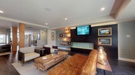 This single-floor amenity space offers a thoughtfully designed environment that blends modern style with practical functionality. The centerpiece is an expansive lounge area featuring rich, dark hardwood flooring, a bold accent wall with a wall-mounted flat-screen TV, a sleek linear gas fireplace, and floating wood shelves with ambient pendant lighting. A generously sized tufted leather sofa anchors the space, complemented by accent chairs and a reclaimed wood coffee table atop a striped area rug. Large windows along one wall flood the lounge with natural light and provide views of lush greenery outdoors, enhancing the open and airy atmosphere. A well-appointed kitchenette serves the common area, outfitted with dark cabinetry, white quartz countertops, a subway tile backsplash, stainless steel appliances including a microwave and beverage refrigerator, a utility sink, and open wood shelving illuminated by industrial-style sconce lighting. The fitness center is comprehensively equipped with treadmills, elliptical machines, stair climbers, and Cybex strength-training equipment, all set on rubberized flooring with natural light streaming through windows. Together, these spaces create a polished, functional environment suited for both relaxation and an active lifestyle.