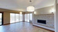 This single-floor space offers a thoughtfully designed open-concept living area that combines modern aesthetics with functional elegance. The main living space is anchored by a striking stone-clad fireplace wall, featuring a sleek, linear electric or gas fireplace set against a textured natural stone surround that extends from floor to ceiling, serving as a bold focal point in the room.

Large sliding glass doors line the perimeter of the living area, flooding the space with abundant natural light and offering expansive views of the surrounding outdoor environment. Sheer roller shades provide privacy and light control while maintaining the bright, airy ambiance of the room.

The flooring throughout is composed of large-format neutral-toned tiles, lending a clean and sophisticated character to the space. Warm wood base trim provides a contrasting accent that adds visual depth and warmth.

Overhead, a combination of pendant lighting fixtures enhances the room's modern character — a large drum-shade pendant with a polished chrome fixture takes center stage, complemented by a decorative globe-style pendant in a secondary area of the room. Recessed wall sconces add subtle ambient lighting along the fireplace wall.

The neutral color palette of warm beige and white tones throughout the walls and ceiling creates a calm, inviting atmosphere that is both versatile and timeless.