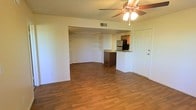 This single-floor space features an open-concept layout that seamlessly connects the living area to the kitchen. The living area is finished with warm hardwood-style flooring that extends throughout, creating a cohesive and inviting atmosphere. A ceiling fan with a built-in light fixture is centrally positioned overhead, providing both air circulation and ambient lighting.

A defining architectural detail is the arched passageway that transitions the living space into an adjoining area, where built-in wall shelving offers practical storage and display options. The walls are finished in a clean, neutral tone that enhances the sense of openness throughout.

The kitchen is accessible via a peninsula-style counter with granite countertops, providing both a workspace and a natural divider between the cooking and living areas. Dark wood cabinetry pairs with stainless steel appliances, including a refrigerator and what appears to be a built-in microwave. A HVAC vent is positioned near the kitchen area for climate control. The front entry door is located adjacent to the kitchen, providing a straightforward and functional entry point into the space.