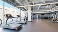 Located at 1980 N Milwaukee Ave in Chicago's Bucktown neighborhood, this contemporary fitness center offers a well-equipped, single-floor space designed for a full range of workout experiences. The facility features an open floor plan with high exposed ceilings that reveal industrial ductwork and structural elements, lending the space a modern, urban aesthetic. Linear LED lighting runs throughout, ensuring bright, even illumination across the entire floor.

Large floor-to-ceiling windows line the perimeter, flooding the space with abundant natural light and providing views of the surrounding streetscape. The durable rubber flooring throughout supports both cardio and strength training activities. Cardio equipment includes Star Trac treadmills, stationary bikes, ellipticals, and rowing machines, while the strength training area offers a cable machine, adjustable benches, plyometric boxes, medicine balls, and a full rack of dumbbells. Wall-mounted flat-screen TVs are positioned throughout for entertainment during workouts.

A mirrored accent wall expands the sense of space in the strength training zone, and a separate, slightly partitioned area with a yellow accent shelving unit and motivational wall text adds a distinct, personality-driven corner to the facility. The overall ambiance is clean, energetic, and functional, making this fitness center a well-rounded destination for residents and members alike.