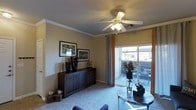 This single-floor residence features a well-appointed living room that serves as the central gathering space. The room is finished with plush wall-to-wall carpeting and warm, neutral-toned walls that create an inviting atmosphere. Crown molding runs along the perimeter of the ceiling, adding a refined architectural detail, while a ceiling fan with an integrated light fixture provides both comfort and illumination.

A prominent sliding glass door floods the space with natural light and provides direct access to an outdoor patio area, where additional seating is visible, extending the usable living space outdoors. Sheer curtains frame the door, allowing light to filter through while offering privacy when desired.

The living room is thoughtfully arranged, featuring a dark wood credenza along one wall, adorned with decorative vases and framed artwork above it. A glass-topped coffee table anchors the seating area, complementing the overall aesthetic. The combination of natural light, tasteful décor, and functional design elements gives this living space a comfortable and well-balanced ambiance.