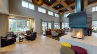 The Sugarloaf Grove Clubhouse in Lawrenceville, Georgia is a thoughtfully designed community gathering space that combines contemporary style with warm, inviting details. The centerpiece of the main hall is a dramatic multi-sided fireplace suspended from the soaring ceiling, surrounded by large-format tile flooring that extends throughout the open-plan interior. Coffered ceilings with rich wood accents and oversized black-and-gold pendant lights lend an upscale, modern aesthetic to the expansive common area. Floor-to-ceiling windows and multiple sets of glass doors flood the space with natural light while offering views of the outdoor pool and surrounding community grounds. The lounge area features comfortable, stylishly upholstered seating arrangements with accent ottomans in bold colors, creating inviting spots for casual gatherings. A long wooden reception and workstation bar with bright accent seating serves as the leasing and business hub, accompanied by a private conference room enclosed in glass. Additional seating nooks with large windows provide quiet retreats overlooking the pool area. Multiple entry points connect seamlessly to the outdoor surroundings, reinforcing the clubhouse's role as a social and functional anchor for the Sugarloaf Grove community.