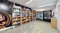 This compact convenience retail space is thoughtfully designed to serve the needs of an active clientele, positioned directly adjacent to a fitness center accessible through full-length glass-paneled doors. The single-room layout makes efficient use of its footprint, with multiple rows of light wood open shelving units lining the walls and stocked with a wide variety of goods — including snack foods, candy, chips, household cleaning supplies, paper products, and health and wellness items. Bold, large-format athletic imagery adorns the back wall, reinforcing the space's connection to the adjoining fitness facility and adding a dynamic visual element to the retail environment. Along one wall, two glass-door refrigeration units display an array of chilled beverages, from bottled water to energy drinks and sodas. A small checkout station with a tablet-based point-of-sale system is conveniently positioned near the refrigerators, alongside a set of large louvered storage cabinet doors that offer additional back-of-house storage. Overhead, recessed linear LED lighting provides bright, even illumination throughout. The floor features a patterned commercial carpet tile in grey and yellow-green tones, adding texture and color to the space. Overall, the retail area is clean, modern, and highly functional.