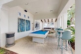 Located at 270 Courtney Lakes Cir in West Palm Beach, Florida, this single-story clubhouse facility offers a thoughtfully designed resort-style amenity space that seamlessly blends indoor and outdoor living. The interior game and lounge area features soaring vaulted ceilings accented by exposed dark wood beams, crisp white walls, and herringbone-patterned stone tile flooring throughout. A standout white pool table with a vivid blue felt surface anchors the game room, complemented by a trio of large tropical botanical art panels and decorative geometric window accents that allow natural light to filter in. A patterned encaustic tile feature wall frames a wall-mounted flat-screen television, creating a comfortable media lounge with natural wood-framed seating and tropical-print cushions. Bar-height tables and teal chairs line the perimeter, offering additional casual seating. The space transitions seamlessly to an open-air covered terrace supported by structural columns and exposed beam ceilings, where neutral-toned lounge furniture is arranged around a low white coffee table atop a lush tropical-print area rug. An outdoor wet bar with dark wood cabinetry overlooks a resort-style pool lined with lounge chairs and shaded by towering palm trees. The lushly landscaped courtyard entry, filled with fan palms and tropical plantings, enhances the vibrant Florida ambiance throughout.