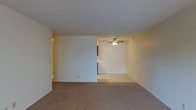 This single-story space features an open-concept layout that creates a sense of spaciousness and easy flow between areas. The main living area is finished with wall-to-wall neutral-toned carpet, providing a comfortable and cohesive feel underfoot. Walls are painted in a clean, off-white hue that reflects light evenly throughout the space.

A partial dividing wall separates the main living zone from an adjoining area, adding subtle definition without fully closing off the floor plan. The adjoining section is equipped with a ceiling fan fitted with integrated lighting, offering both air circulation and ambient illumination. The textured ceiling adds a classic residential character consistent throughout the space.

A hallway visible toward the back suggests access to additional rooms or utility areas, hinting at a functional layout designed for practical everyday use. The overall design is straightforward and neutral, offering a blank canvas adaptable to a variety of furnishing styles. The consistent use of light tones across walls, ceilings, and flooring contributes to a bright, airy ambiance throughout the space.