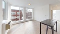 Located at 413 Grand, this single-floor unit presents a clean, modern design characterized by light wood-style flooring and a crisp white color palette throughout. The open-concept layout seamlessly connects the kitchen and living areas, creating a cohesive and functional living space.

The kitchen is well-appointed with white flat-front cabinetry extending to the ceiling, light-toned countertops, and a full suite of stainless steel appliances, including an electric range, built-in microwave, dishwasher, and refrigerator. A dark-topped island or bar table anchors the kitchen, providing additional prep and dining space. Track lighting overhead ensures excellent task illumination.

The living area is generously proportioned and bathed in natural light from a large bank of floor-to-ceiling windows that frame views of the surrounding neighborhood. The bright, open atmosphere is enhanced by the continuity of light flooring and neutral white walls throughout.

The bathroom maintains the unit's minimalist aesthetic with white cabinetry, a clean vanity with ample storage, and a full soaking tub with a shower. Wood-style flooring carries through into this space, adding warmth to the otherwise clean, functional design.