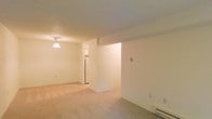 This single-floor space presents a straightforward, functional layout characterized by clean lines and a neutral color palette throughout. The main living area is finished with light-colored carpeting that extends across the open floor plan, contributing to a warm and understated ambiance. Walls are painted in soft, off-white tones, creating a blank canvas well-suited for personalization.

A pendant light fixture is visible near the transition point between the living area and the kitchen, providing focused overhead lighting. The kitchen can be glimpsed beyond a partial wall divider, where a refrigerator is visible, suggesting a compact but practical culinary space. The partial wall creates a subtle separation between the living and kitchen areas without fully closing off the floor plan.

A baseboard heating unit lines one wall, indicating a dedicated heating system for the space. The textured ceiling adds a modest architectural detail, while multiple electrical outlets are positioned along the walls for convenience. Overall, the space offers a simple, move-in-ready environment with a cohesive, neutral design that prioritizes practicality and ease of use.