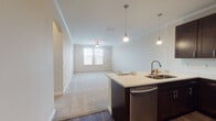This single-floor residence offers a thoughtfully designed open-concept layout that seamlessly connects the kitchen and living area. The kitchen is a standout feature, outfitted with rich dark-wood cabinetry extending to the ceiling, stainless steel appliances including a refrigerator, range, and microwave, and a light-colored countertop island with a double sink. Stylish pendant lights hang above the island, which also provides casual seating with bar stools. The kitchen transitions smoothly into the carpeted living area, which benefits from natural light streaming through windows fitted with blinds, and is finished with crown molding and a ceiling fan. An arched doorway adds an architectural detail that enhances the flow between spaces. The bedroom features plush carpet flooring, neutral walls, and dual windows that bring in ample natural light, along with access to a walk-in closet. The bathroom is well-equipped with a dark-wood vanity, white countertop, large mirror with overhead lighting, and a tub-shower combination. Throughout the residence, wood-look flooring in the kitchen and bathroom contrasts elegantly with the soft carpet in the living and sleeping areas, creating a cohesive and comfortable atmosphere.