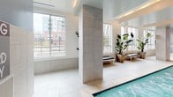 Located at Uptown 550 Stonewall in Charlotte, North Carolina, this indoor pool and spa facility is thoughtfully designed to provide a refined amenity space within an urban setting. The single-level space showcases a clean, contemporary aesthetic defined by large-format light-toned floor tiles, floor-to-ceiling tiled columns, and smooth wall surfaces finished in coordinating neutral tones.

The pool runs the length of the space and is framed by wide walkways on either side, providing ample room for movement. Tall windows line the perimeter walls, flooding the interior with abundant natural light while offering views of Charlotte's surrounding cityscape and adjacent outdoor terrace areas. The outdoor views give the enclosed pool area an open, airy quality despite its interior setting.

Pendant light fixtures hang from the ceiling, supplementing the natural light and contributing to the polished atmosphere. Potted tropical plants are positioned throughout the space, introducing a natural element that softens the overall design. Sleek benches provide seating areas along the poolside. The entryway features a branded welcome mat identifying the Uptown 550 Stonewall property, reinforcing the space's identity as a signature amenity within this Charlotte residential community.