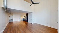 Located at 780 S 52nd St in the West Philadelphia neighborhood of Philadelphia, PA, this two-floor space offers a well-designed layout across a generously sized main level and a secondary upper level. The main floor commands attention with its dramatically soaring ceilings, accentuated by a striking black ceiling fan that reinforces the industrial-modern aesthetic. Rich hardwood flooring runs throughout, providing warmth against the crisp white walls. Large floor-to-ceiling glass panels on the entry side flood the space with natural light and provide views of the surrounding neighborhood. The open-concept living area flows seamlessly into a compact kitchen fitted with stainless steel appliances, including a full-size refrigerator, and dark cabinetry that adds contrast to the bright interior. Recessed lighting in the kitchen zone ensures functional illumination, while exposed ductwork adds an industrial character to the upper living area. The bathroom is a standout feature, finished with bold geometric patterned floor tile in charcoal and gold, a frameless glass-enclosed shower, a floating wood-accented vanity, and black hardware throughout. The upper level provides additional versatile living space accessible via an interior staircase, completing this thoughtfully designed multi-level property.