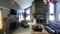 This single-floor space offers an impressive open-concept layout defined by dramatic vaulted ceilings that create an expansive, airy atmosphere. The main lounge area is anchored by a stone-faced fireplace, providing a warm focal point complemented by large windows dressed in sheer curtains that invite abundant natural light. Comfortable seating arrangements, including plush armchairs with ottomans and a sofa, surround a dark wood coffee table, creating a welcoming and well-appointed gathering space. The walls are finished in a deep, sophisticated gray tone, lending a modern and refined character throughout. A large flat-screen television is mounted above the fireplace, adding a contemporary touch. An elegant chandelier overhead adds a decorative accent to the room. Transitioning seamlessly from the lounge, a bar or kitchenette area features a raised counter with bar-height seating, overhead cabinetry, and what appears to be a mirrored backsplash that reflects light and enhances the sense of depth. Framed artwork adorns the walls, contributing to the curated, stylish ambiance. A tall indoor tree adds a natural, organic element to the space. Overall, this space balances comfort and sophistication, making it well-suited for social gatherings or everyday use.