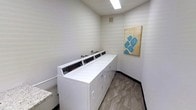 This single-floor laundry facility is designed with both functionality and aesthetics in mind. The room features a row of modern, commercial-grade washing machines aligned neatly along one wall, offering ample capacity for laundry needs. The machines are finished in crisp white, complementing the clean, neutral-toned walls adorned with subtle horizontal striping that adds a touch of visual interest without overwhelming the space. Overhead, a recessed fluorescent light fixture provides bright, even illumination throughout the room, ensuring a well-lit environment for users. The flooring consists of durable, marble-patterned tiles in warm gray tones, lending an upscale feel to an otherwise utilitarian space. A countertop surface is visible to the side, offering a convenient area for folding or sorting laundry. A decorative canvas artwork featuring an abstract blue design on a warm background adds a welcoming, personal touch to the room. A ventilation grille is also present near the ceiling, supporting proper air circulation. Overall, the space strikes a balance between practicality and cleanliness, presenting a tidy and organized laundry room suitable for shared or communal use.
