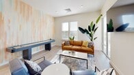 Located at 1601 Overbrook Rd in Richmond, Virginia, this thoughtfully designed amenity space offers residents a vibrant and functional environment for both socializing and focused gatherings. The interior lounge area features light hardwood-style flooring, recessed lighting, and an abundance of natural light streaming through large windows. A striking watercolor-inspired accent wall in soft peach and teal tones serves as a defining architectural detail, anchoring both the lounge and an adjoining private meeting room. The lounge is furnished with a warm cognac leather sofa, coordinating accent chairs, a sleek coffee table, and a wall-mounted flat-screen television, creating an inviting space for casual gatherings. A shuffleboard table adds a recreational element to the room. The private meeting area features a natural wood conference table with contemporary black chairs, ideal for small group collaboration. Lush tropical plants throughout the interior enhance the fresh, modern atmosphere. Through floor-to-ceiling glass doors, the space opens onto a generously sized outdoor rooftop terrace. The terrace is appointed with vibrant orange seating, outdoor dining tables, and a built-in grilling station, all set beneath open skies with expansive views of the surrounding Richmond cityscape.