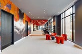 Located at 501 NW Seventh Ave in Fort Lauderdale, FL, this contemporary fitness center delivers a comprehensive, high-design workout environment across a single, expansive floor. The space is defined by its bold aesthetic, featuring striking red perforated ceiling panels with integrated linear lighting that run the length of the main exercise floor, set against dark charcoal walls and durable light-toned flooring. Floor-to-ceiling windows line the perimeter, flooding the interior with natural light and offering open views of the surrounding greenery and streetscape. The main workout hall is generously proportioned, accommodating a full suite of cardio equipment — including treadmills, stair climbers, ellipticals, and stationary bikes — as well as a dedicated strength training zone with cable machines, weight benches, and a free weights rack. A separate spin studio features hardwood flooring, floor-to-ceiling mirrors framed in teal, built-in storage cubbies, and its own set of cycling bikes. An additional mirrored training room with teal accent walls and hardwood floors provides space for functional training, with kettlebells, medicine balls, and resistance equipment readily available. A welcoming lounge area near the entrance features bold red modern seating and large-scale abstract mural art. Two bathrooms and a closet round out the facility's amenities.
