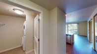 This single-floor residence offers a thoughtfully designed layout that balances functionality and comfort. The open-concept living and kitchen area serves as the heart of the home, featuring rich hardwood-style flooring, a spacious center island with a double sink and generous counter space, and warm light wood cabinetry that extends to the ceiling for ample storage. Stainless steel appliances — including a refrigerator, range, and built-in microwave — complete the kitchen, while pendant and track lighting add a modern, welcoming ambiance. The living area is bright and generously proportioned, with sliding glass doors opening to a private balcony that brings in natural light and pleasant outdoor views of surrounding greenery. The bedroom is carpeted and tranquil, featuring a large picture window framing lush treetop and landscape views, along with a wall-mounted climate control unit for year-round comfort. The bathroom is clean and practical, offering a wood-toned vanity with a dark countertop, a large mirror, and grab bars for accessibility. A dedicated utility closet houses a stacked washer and dryer, providing convenient in-unit laundry. Throughout the space, neutral tones and clean lines create a calm, move-in-ready atmosphere suited to everyday living.