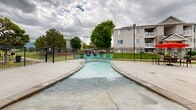 Located at 1801 W 4650 S in Roy, Utah, this outdoor amenity area serves as a central gathering space designed for relaxation and recreation. The centerpiece is a generously sized swimming pool with crystal-clear turquoise water, surrounded by a wide concrete pool deck furnished with lounge chairs and bold red shade umbrellas, offering ample space for sunbathing and socializing. Adjacent to the main pool is a shallow wading pool, ideal for younger guests, featuring vibrant teal Adirondack chairs positioned along its gentle, curved edges. A hot tub with stone accents provides an additional relaxation option and is conveniently situated near a dedicated pool house facility. The pool house structure features a low-pitched roof with stone detailing and houses support amenities for the pool area. The entire complex is enclosed by a sturdy black iron fence, providing security and defined boundaries. Mature trees are scattered throughout the surrounding grounds, adding greenery and natural shade. The multi-story residential buildings visible in the background feature balconies that overlook the amenity area. The well-maintained concrete hardscape throughout the space ensures easy movement between the various aquatic features, creating a cohesive and functional outdoor environment.