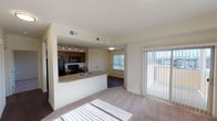 This single-floor residential unit offers a thoughtfully designed open-concept layout that seamlessly connects the kitchen and living areas. The kitchen is equipped with dark-stained wood cabinetry, black appliances including a refrigerator, electric range, and over-the-range microwave, and is complemented by a peninsula with a sink that provides additional counter space and serves as a natural divider between the kitchen and living room. Track lighting illuminates the kitchen, while hardwood-style flooring adds a warm, polished finish. Adjacent to the kitchen, a spacious closet with louvered double doors offers generous storage. The living area features plush carpeting and is bathed in natural light streaming through large sliding glass doors that open to a private balcony, offering views of the surrounding area. Vertical blinds throughout allow for flexible light control. The unit includes multiple bedrooms, each carpeted and naturally lit through windows fitted with vertical blinds, providing a bright and comfortable atmosphere. The full bathroom is clean and functional, featuring a full-size tub and shower combination with chrome fixtures. The overall color palette of soft neutral tones throughout creates a cohesive, airy ambiance, making this a versatile and comfortable living space.