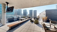 Located in Chicago's vibrant West Loop neighborhood at 123 N Desplaines, this expansive rooftop amenity level offers an impressive array of outdoor features across a generously sized single floor. The centerpiece of the space is a large covered pool surrounded by rows of lounge chairs, ideal for relaxation while taking in panoramic views of the Chicago skyline. Sleek glass railings line the perimeter, ensuring unobstructed sightlines of the surrounding cityscape, including notable high-rises and iconic towers.

The rooftop is divided into several well-appointed zones. A partially covered lounge area features a concrete overhead canopy, providing shade over clusters of stylish seating arrangements, including woven accent chairs, benches with warm wood accents, and dining tables suited for group gatherings. Multiple outdoor grills are positioned nearby, making this area well-equipped for entertaining.

A glass-enclosed interior section connects the outdoor spaces and is accented with lush vertical greenery, offering a bright, climate-controlled retreat. Additional open-air sun decks extend around the pool area, lined with chaise lounges bathed in natural light. A staircase and a bathroom round out the functional amenities. The overall design blends contemporary materials — glass, concrete, and tile — with a clean, modern aesthetic befitting its urban Chicago setting.