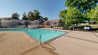 Located at 800 Alexander Rd in Cayce, South Carolina, this outdoor pool area offers a well-appointed recreational retreat designed for relaxation and leisure. The centerpiece is a generously sized swimming pool with clear turquoise water, bordered by a broad concrete sun deck finished with brick accent inlays. Multiple lounge chairs are positioned throughout the deck, providing ample space for sunbathing under the open South Carolina sky.

A distinctive covered pavilion anchors one end of the pool area, featuring a standing-seam metal roof supported by white-painted columns and complemented by white brick walls with curved detailing. Steps lead up to an elevated terrace, where shaded outdoor seating with a large umbrella creates a comfortable gathering spot. A wood pergola with additional seating adds another layer of character to the space.

The pool area is enclosed by black iron fencing and is framed by mature trees that provide natural shade and privacy. Neatly trimmed hedges and ornamental plantings enhance the landscaped surroundings, contributing to an inviting, park-like atmosphere. The adjacent multi-story residential buildings, painted in blue and gray, are visible in the background, contextualizing this amenity as a shared outdoor facility within a well-maintained residential community.