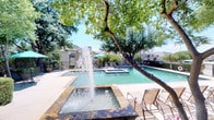 Located in Lewisville, Texas, this outdoor recreational space centers around a generously sized swimming pool with clear, blue-green water and classic brick-edged coping. An integrated spa sits directly adjacent to the main pool, offering a dedicated relaxation zone with water jets. A standout decorative fountain feature adds visual interest and an inviting ambiance to the pool deck.

The surrounding pool deck is finished with a light-colored surface and is well-equipped with multiple lounge chairs positioned to take advantage of the Texas sunshine. Several teal market umbrellas are strategically placed throughout the area, providing shaded spots for relaxing. Mature, leafy trees border the space, offering additional natural shade and a lush, landscaped feel that softens the hardscape elements.

Neatly trimmed hedges and ornamental plantings line portions of the perimeter, contributing to a well-maintained, resort-like atmosphere. A clubhouse-style building with a covered porch and additional seating areas is visible along one side of the pool area, suggesting on-site amenity support. The space is enclosed by a black iron perimeter fence, ensuring privacy and security. Overall, this outdoor area delivers a comfortable and attractive setting ideal for leisure and recreation under open Texas skies.